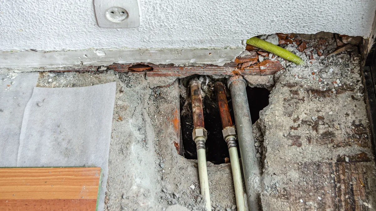 Aufgestemmter Fußboden an einer weißen Wand: Im grauen Estrich ist ein Loch geöffnet, darin verlaufen zwei rostbraune Rohre, die über Verschraubungen in helle Kunststoffrohre übergehen, daneben ein graues Rohr; rechts steckt ein gelbgrünes Leerrohr im aufgebrochenen Wandsockel aus Ziegeln, oben links sitzt eine Steckdose, links liegen eine helle Unterlagsbahn und ein Stück Laminat